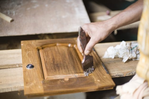 Close-up of a hand applying a wood finish to a cabinet panel in a workshop